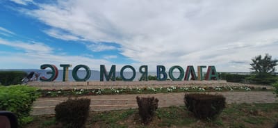 The image captures a wide-angle view of a large sign that reads "ЭТО МОЯ ВОЛГА" (This is My Volga) situated along a waterfront. The letters of the sign are colorful, with each letter having a green border and a different colored interior, such as yellow, red, and blue. The sign is placed on a concrete base adorned with a line of flowers in shades of white, pink, and yellow, with some darker red flowers interspersed.
To the left of the sign, a bicycle with a basket is leaning against a green shrubbery. The background features a body of water, likely the Volga River, with a distant shoreline and a partly cloudy sky. The foreground consists of a paved walkway made of brick-like stones, bordered by grass and more shrubs. To the far right, a mature tree stands near the water's edge.
The scene is set in the city of Rovnoye, Russia, specifically at its waterfront park, a common recreational area with public art and natural landscaping. The overall atmosphere is serene and picturesque, suggesting a pleasant day by the river.