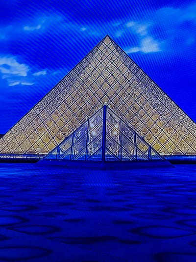 The scene depicts a large, illuminated glass pyramid under a deep blue sky with minimal cloud cover. The primary pyramid structure is composed of numerous small, uniformly arranged diamond-shaped glass panes, emitting a warm, golden-yellow light against the dark blue backdrop. In front of the main pyramid, a smaller, multi-faceted glass pyramidal entrance structure is visible, also constructed from clear glass panes and exhibiting internal structural elements. The ground surface extending from the base of the structures is uniformly dark blue, presenting subtle, irregularly shaped lighter blue patterns or reflections across its surface. No individuals or specific activities are observable. This scene is located in the city of Madzherito, Bulgaria.