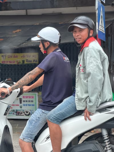 Two men are sitting on a white motorcycle in what appears to be a city street in Quảng Trị Ward, Vietnam. The man in the front is wearing a dark blue t-shirt and denim shorts, with tattoos on his left arm and a silver bracelet on his left wrist. He is wearing a white helmet. The man behind him is wearing a light gray jacket with a red collar over a red shirt, and a black helmet. He has his left hand resting on the seat of the motorcycle. In the background, there is a building with signs in Vietnamese and a blue traffic sign with a white arrow. The lighting suggests it is daytime.