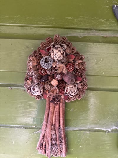 A decorative, tree-shaped natural arrangement is vertically mounted against an olive-green horizontal wooden paneled surface in the city of Stiperstones, United Kingdom. The item measures approximately 40 cm in height and consists of a dense, rounded top section composed of various dried natural elements, including numerous small pine cones, larger textured seed pods, and stylized flower-like components, some appearing silvery and others reddish-brown. The lower section forms a bundled "trunk" constructed from vertical cinnamon sticks or similar reddish-brown wood bundles, with several star anise pods embedded near the top of this section. The entire arrangement and the adjacent green paneling exhibit visible accumulations of fine dust and white cobwebs, indicative of prolonged exposure to ambient conditions without recent cleaning. No individuals, actions, or direct interactions are depicted.