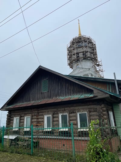 In the city of Kstínino, Russia, a wooden log building with a dark, weathered facade stands with a prominent, cupola-topped structure undergoing renovation behind it. The main building features several white-framed windows and a steeply pitched, dark wooden roof. A green metal fence encloses the property, with overgrown vegetation visible along the fence line and at the base of the building. Power lines stretch across the overcast sky above the scene. The cupola is partially encased in scaffolding, indicating ongoing construction or repair work. The overall impression is one of a rural or semi-rural setting with a blend of aged architecture and active restoration.