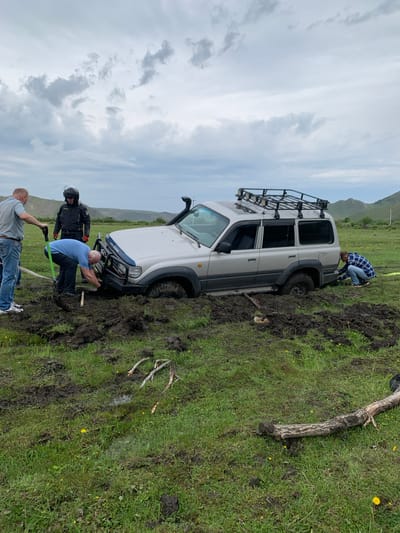 In the city of Khurai-Nur, Russia, a silver SUV is deeply stuck in mud in a grassy, uneven field under a cloudy sky. Four individuals are present, seemingly attempting to free the vehicle. One person, wearing a helmet and jacket, stands by the front of the SUV. Another person in a blue shirt is using a shovel to dig around the front bumper. A third person, in a plaid shirt, is kneeling at the rear of the vehicle, near a yellow tow strap. A fourth person, wearing a plaid shirt and jeans, stands to the left of the SUV, also holding a shovel. The ground is a mix of mud and grass, with scattered yellow wildflowers. Several tree branches lie on the ground in the foreground. Hills are visible in the background.