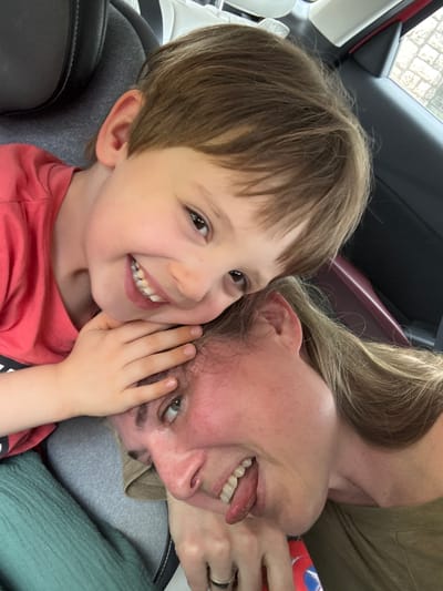 A selfie shows a smiling young boy with brown hair and a red shirt seated in a car seat, cheek-to-cheek with a fair-skinned adult. The adult, with blonde hair and wearing a green shirt, is also smiling and sticking out their tongue. The car's interior features a black and gray car seat, a burgundy trim, and a white window frame. The location is identified as Meljine, Montenegro.