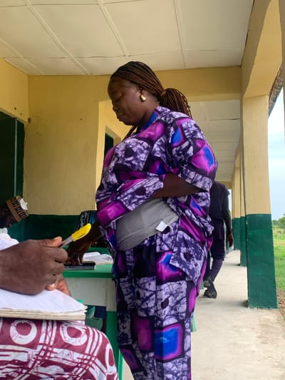 A woman in a purple patterned garment leans forward, appearing to examine or interact with something out of frame, while another person holds a notebook and pen in the foreground. The scene is set in what appears to be a corridor or covered walkway, with yellow and white painted walls. Another individual sits in the background, legs visible. The location is Tabu, Nigeria, based on the context provided.