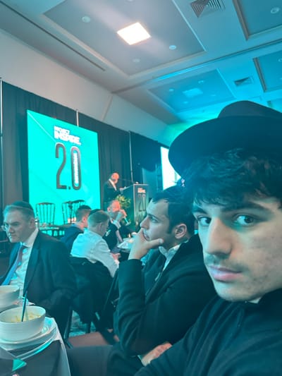 A selfie captures two men at a convention. The foreground man wears a hat and looks at the camera. Behind him, another man contemplates, hand to chin. A large screen displays "Inside 2.0 Convention". Several other attendees are seated, facing a stage where someone is speaking into a microphone. Plates and bowls sit on a table in the bottom right corner. Ceiling lights and air vents are visible above. The image has a slight blue tint.