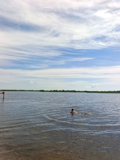 A person is swimming in the calm, dark water of a lake. The sky is blue with wispy white clouds. Trees and a distant shore are visible in the background. There is a wooden post to the left of the frame, sticking out of the water. The scene appears to be in Vyazovka, Russia.