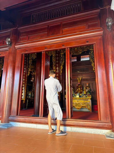 A medium shot, taken from a slightly low angle, captures a man standing at the threshold of an ornate, wooden structure, possibly a temple or historical building. The image is oriented such that the top of the building is on the left and the base is on the right. The man is dressed in a light-colored shirt and shorts, wearing dark-colored sandals. He appears to be looking into the building, his posture suggesting reverence or contemplation.

The building itself is constructed from dark wood, featuring intricate carvings and details. The interior is richly decorated with gilded ornaments and statues, creating a sense of opulence and history. The floor outside the building is tiled in a warm, terracotta color.

Based on the ornate architectural style and the presence of religious iconography, the location is likely in Diên Lạc, Vietnam, known for its historical and religious sites. The man's attire and behavior suggest he may be a tourist or a local visiting the site for prayer or cultural enrichment. The overall impression is one of cultural heritage and spiritual significance.
