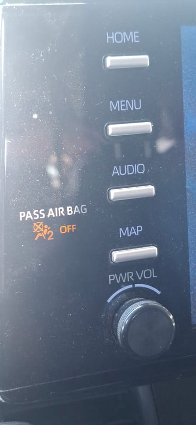 A car's infotainment system display is visible, showing several buttons and a warning light. The buttons are labeled "HOME," "MENU," "AUDIO," and "MAP." Below the "PASS AIR BAG" text, there is an illuminated orange symbol indicating the passenger airbag is OFF. A large, circular knob labeled "PWR VOL" is at the bottom of the display, likely for power and volume control. The dashboard is dark and has a matte finish, with specks of dust visible on the screen. The background is blurred, suggesting the image was taken from inside a vehicle. The specific city in Israel is not discernible from the image.