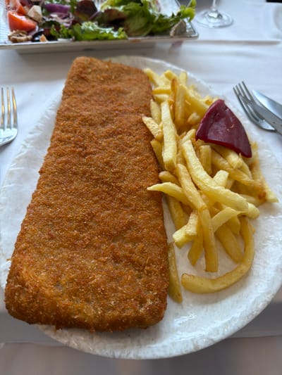 The image displays a large, breaded and fried cutlet, characterized by its golden-brown, textured crust, positioned on the left side of a white, oval-shaped plate. To the right of the cutlet, a mound of golden-yellow french fries is present, garnished with a single, dark red roasted pepper. The food rests on a sheet of white paper or absorbent material on the plate. A silver four-tine fork is visible on the white tablecloth to the left of the plate, and a silver fork and knife are partially visible to the right. In the blurred background, a rectangular dish containing a green salad with tomatoes and red onions, along with the stem and base of a clear wine glass, are discernible. The scene depicts a prepared meal on a table in a restaurant located in Colunga, Spain. No individuals or active interactions are visible.