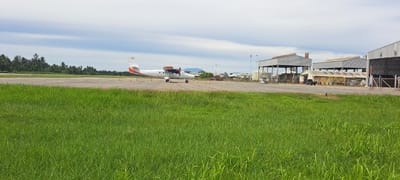 A twin-engine airplane is parked on the tarmac of a small airport in Keudeblangjruen, Indonesia. The plane, marked with "PEGASUS" on its side and the registration "PK-TUJ," is white with orange and black accents. It is positioned on a paved runway, with several orange traffic cones visible nearby. In the background, there are a few low-rise buildings, some appearing to be hangars or storage facilities, with corrugated metal roofs. A dark-colored pickup truck is parked in front of one of the buildings. Beyond the airport structures, a dense line of palm trees and other tropical vegetation is visible, suggesting a humid, warm climate. The sky is overcast with scattered clouds. The foreground is dominated by lush, green grass.