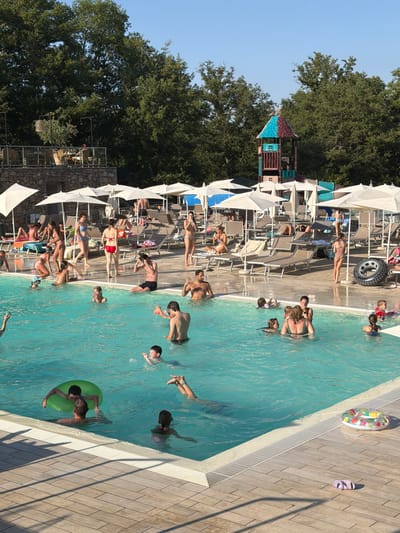 The image depicts a crowded outdoor swimming pool area on a sunny day. The pool is filled with people of various ages swimming, playing, and relaxing. Several individuals are partially submerged in the turquoise water, some with inflatable toys. Around the pool, lounge chairs are set up under white umbrellas, with some people sunbathing or sitting on them. In the background, trees form a dense green backdrop, and a colorful playground structure is visible. The surrounding area features a paved pathway with a wooden deck pattern and a stone wall. The scene suggests a resort or water park environment in Cavriglia, Italy, where people are enjoying recreational activities.