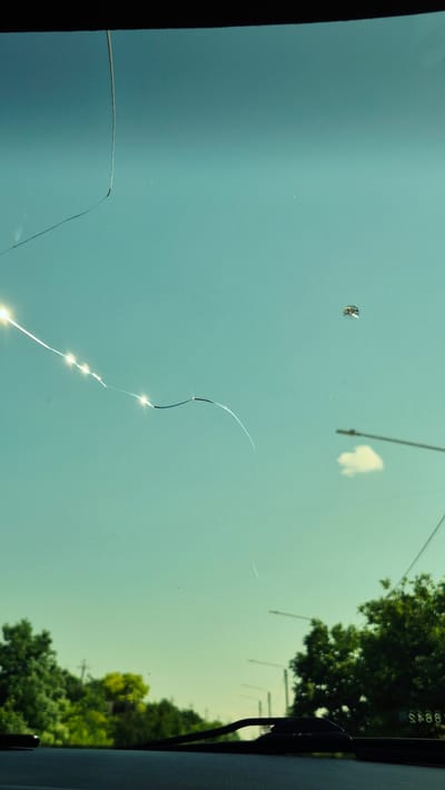 The image captures a view through a cracked car windshield. The sky is a clear, pale blue with a gradient. A prominent crack runs diagonally from the upper left corner, with several bright glints of light reflecting off its fractured surface, indicating direct sunlight. Further to the right, a small, spiderweb-like impact mark is visible on the glass. In the distance, a single, small white cloud is present. Below the sky, a line of green trees forms the horizon, with some lighter green foliage interspersed. Streetlights are visible amongst the trees, and in the bottom right corner, a portion of a car's dashboard and windshield wipers are in focus. A partially obscured sign with reversed lettering is visible on the right side of the image. The overall impression is that of a sunny day, viewed from inside a vehicle, with the damage to the windshield being the focal point. The location of Mihailovca, Moldova, is not depicted within the image itself; this information is external to the visual content.