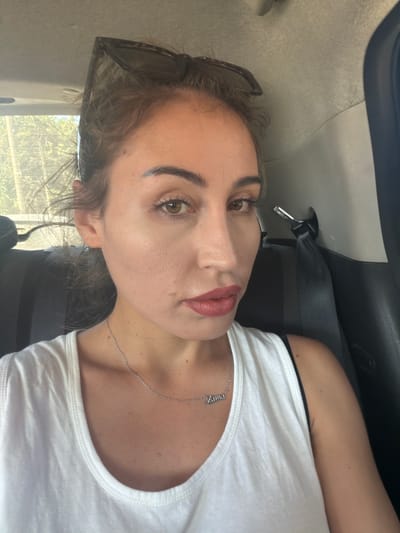 A woman is seated in the back of a vehicle, possibly a car, with a seatbelt fastened behind her. She is looking towards the camera, and her face is partially illuminated by natural light coming from an unseen window. Her hair is pulled back, and she is wearing sunglasses on top of her head. She has on a white tank top and a silver necklace with a nameplate. The background shows the interior of the car, including a headrest and upholstery, as well as a blurred view of trees and foliage through a window, suggesting an outdoor environment. The scene appears to be located in Dromolaxia - Meneou Municipality, Cyprus, given the context provided.