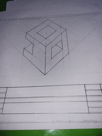 The image displays a white sheet of paper laid flat on a distinct green surface. The paper itself features a technical drawing. In the upper half, an isometric projection of a three-dimensional object is rendered in black lines, depicting a hollowed, cube-like structure with a central rectangular cut-out on its top surface and two open, trapezoidal sections on its visible vertical faces. Lighter pencil construction lines, converging towards vanishing points, are visible around this drawing. The lower half of the paper shows several dark horizontal and vertical lines forming multiple rectangular fields, consistent with a title block or layout for additional views. The paper's surface exhibits minor texture and subtle variations in brightness. The background above the paper is an out-of-focus, light gray or off-white surface. The scene is located in the city of Puerto López, Colombia. No people or actions are depicted.