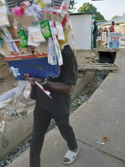 A man carries a blue cooler with assorted goods piled high. He wears black pants, a black shirt, and white sandals, walking along a concrete walkway. A dirty drainage ditch runs alongside the walkway, littered with trash. In the background, people stand near a "SOLAKEM COMMUNICATIONS POS" sign and a small building with a green roof. The scene appears to be outdoors in a busy, possibly urban, area.
