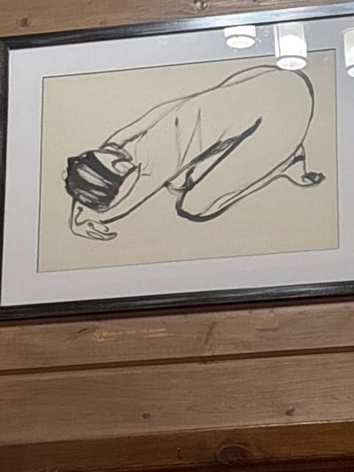 A framed sketch hangs on a wooden wall. The sketch depicts a figure bent over, head down, and reaching for its feet. The lines are bold and suggestive rather than precise. The figure's curves and limbs are defined with thick, black strokes against a light background. The frame is a dark gray color.