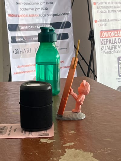 The image shows a close-up of a wooden table with various objects. In the foreground, a black cylindrical speaker is placed on top of a pink receipt. To the right of the speaker, an orange object is propped up by a small, pink figurine that appears to be a stylized bear. Behind these items, a bright green water bottle stands upright. In the background, several posters are visible. One poster on the left has Indonesian text and lists rental terms, including "TENOR DAN TARIF SEWA" and "<30 HARI". Another poster on the right has a red header that reads "LOWONGAN" (vacancies) and "KEPALA O..." (Head of...). A QR code is also visible on this poster. The lighting suggests it is daytime, and the environment appears to be indoors, possibly a shop or office. The presence of receipts, rental information, and job postings suggests a commercial setting in Boja, Indonesia.