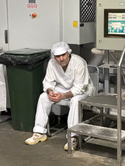 A man in a white uniform and cap sits on a metal chair in what appears to be an industrial setting, possibly a food processing facility. He is wearing white pants and white athletic shoes with yellow soles. His hands are clasped in front of him. To his left is a large green trash bin with a black liner. Behind him, a wall-mounted control panel with a screen displaying various readings is visible. The floor is concrete, and there are metal structures and equipment around the scene. The overall environment suggests a workplace with a focus on hygiene and industrial processes. The specific location in Dedovsk, Russia, is not discernible from the visual information alone.
