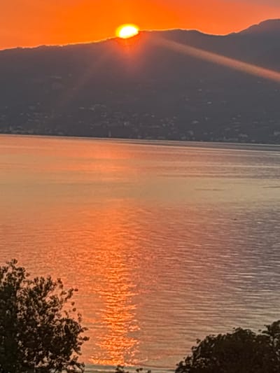 The image depicts a sunset over Lake Garda, Italy. The sun, a brilliant orange orb, is partially obscured by a dark, mountainous silhouette in the background, casting a golden glow across the water. Sunlight streaks through the atmosphere, creating a dramatic beam of light. The lake's surface is calm, reflecting the vibrant colors of the sunset. In the foreground, the dark shapes of trees frame the lower portion of the scene. The overall impression is one of natural beauty and tranquility.