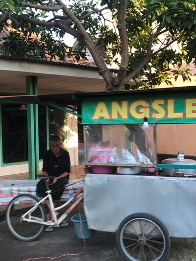 A man wearing glasses and a black polo shirt sits on a bench next to a food cart in Kalidawir, Indonesia. The food cart has a green banner with yellow lettering that reads "ANGSLE". A bicycle is parked in front of the cart. The man is holding a smartphone. A blue bucket is attached to the side of the food cart. In the background, there is a building with a tiled roof and a tree with green leaves. The sun is shining, casting shadows on the ground.