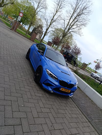 The image captures a bright blue BMW M2 parked on a gray herringbone brick road. The license plate "K-733-XL" is visible. In the background, behind the car, are other vehicles, including a black van with the word "SPAK" and a silver Audi. There are trees, some with green leaves, others with bare branches, as well as brick structures and signage along the street. The location is Emmerich am Rhein, Germany, as indicated by the license plate. The sky is overcast, suggesting a cloudy day.