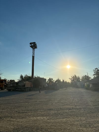 This media file depicts an outdoor scene at what appears to be a construction or industrial site. The sun is low in the sky, suggesting it is either early morning or late afternoon, and it casts a bright glare, creating lens flare effects and a generally hazy atmosphere.

Dominating the left side of the frame is a tall, metallic tower with what looks like equipment or lights at the top. This structure stands against a clear blue sky. Below and to the right of the tower, several vehicles are parked. These include a truck with a concrete mixer drum and what appears to be other heavy machinery or construction equipment. A person is visible standing near one of the trucks.

The ground is covered in gravel or dirt, typical of an outdoor work area. In the background, there are some buildings and trees, indicating a semi-rural or industrial setting. The overall impression is one of a working environment bathed in the warm light of the sun. There is no visible text in the image. The location context provided is Zhirnov, Russia.