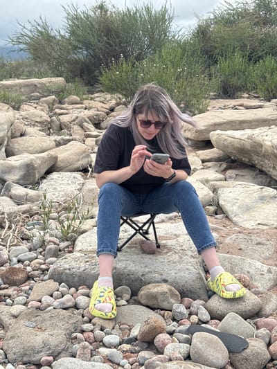 A woman with purple-tinted hair and sunglasses sits on a portable black folding chair on a rocky shore near Tong, Kyrgyzstan. She is wearing a dark t-shirt, blue jeans, and yellow slide sandals with a cartoon pattern. The woman is looking down at her phone, which she holds in her hands. Her phone is a light green color. The background consists of large rocks, smaller pebbles, and sparse green vegetation. The sky is overcast.