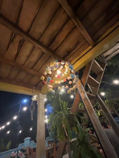 The image captures a whimsical and softly lit outdoor scene, likely at dusk or in the evening, in a tropical setting like Karimunjawa, Indonesia. The primary focus is a decorative, spherical chandelier crafted from colorful pom-poms of various hues including yellow, blue, pink, and red. This light fixture hangs from a rustic wooden structure that suggests an open-air pavilion or a similar architectural element characteristic of island resorts.
The background reveals a complex interplay of natural and man-made elements. Dense green foliage is visible, hinting at the lush tropical environment. Above, wooden beams and rafters form a lattice-like ceiling, through which other lights and the night sky are partially visible. The overall ambiance is relaxed and inviting, suggesting a leisure or dining area.
In the upper right portion of the image, parts of people can be seen, their forms slightly obscured by the lighting and angle. One individual appears to be leaning forward, their face partially visible. These figures contribute to the sense of a social gathering or a place where people are enjoying themselves.
String lights are festooned across the scene, casting a warm glow and accentuating the evening atmosphere. One prominent white column with decorative rope detailing stands to the right of the pom-pom chandelier, further enhancing the decorative and possibly exotic aesthetic of the location. The presence of these lights and the style of the structures strongly indicate a resort, cafe, or restaurant designed to create a magical evening experience. No discernible text is visible in the image.