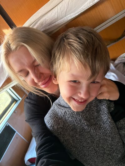 The image depicts an adult female and a young male child in a close-up, head-and-shoulders composition, set in an interior location identified as Bistrice, Montenegro. The female, with blonde hair, is positioned behind and to the left of the child. Her eyes are closed, and a subtle smile is present on her lips. She wears a dark, long-sleeved garment. Her right arm is visible, extended around the child's shoulder area. The child, with light brown hair, is in the foreground, facing the camera with a wide smile, displaying visible teeth. He wears a grey, ribbed knitted sweater. The background consists of light brown wooden paneling. In the upper right, white, crumpled fabric, possibly a curtain or bedding, is visible. A window frame is partially visible in the lower-left background, revealing a blurred exterior with hints of light and natural elements. Below the window, a dark, rectangular metallic vent or grille is present. The interior design suggests a confined space, consistent with a vehicle cabin such as a train or recreational vehicle.