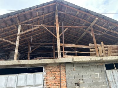 The image depicts a dilapidated structure in Novo Leski, Bulgaria. The lower portion of the building is constructed with a combination of red brick and grey concrete blocks. A large, closed, grey metal gate is visible on the left, and a smaller opening with a partially open metal gate is on the right. Above the brick and concrete wall is a concrete lintel. The upper structure is an open, decaying wooden roof framework, revealing the support beams and roofing structure. There are stacks of tires stored within the building frame. Thin wire runs across the open upper structure. The sky is visible in the background.