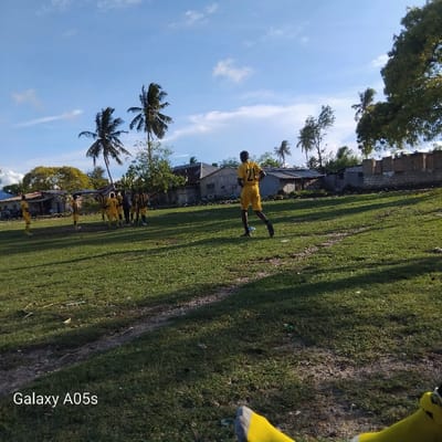 The image shows a soccer field in Nungwi, Tanzania, located at coordinates -5.726 latitude and 39.301 longitude. A group of men in yellow jerseys are playing soccer on a grassy field. The field is surrounded by buildings and trees. The sky is blue and clear. The image is taken from a low angle, with a person's legs and feet visible in the foreground. The "Galaxy A05s" watermark is at the bottom left of the image.
