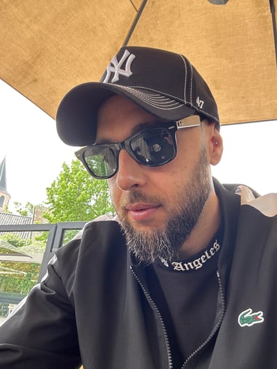 A male individual is depicted in an outdoor setting in Emmen, Netherlands, captured in a close-up, slightly elevated selfie perspective. The individual, an adult, has a dark beard with visible grey hairs concentrated around the chin. He wears a black New York Yankees baseball cap featuring a white "NY" logo on the front and a "47" logo on the right side, along with white decorative stitching. Dark sunglasses, with a silver rectangular accent on the temples, cover his eyes and reflect a person holding a mobile device and a white cup, suggesting the image is a selfie. A small silver stud piercing is present on the upper part of his right ear (helix). He is dressed in a black zip-up jacket, displaying a green Lacoste crocodile emblem with a red mouth on the left chest. A black shirt with white 'Angeles' text in an Old English font is visible beneath the jacket's collar. The scene is shaded by a large, beige patio umbrella. In the background, beyond a glass railing, are green deciduous trees and a brick building with a pointed church spire, viewed under an overcast sky.