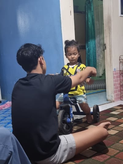 A young child wearing a yellow and black shirt sits on a small blue tricycle in a room with blue walls. An older person wearing a black shirt stands beside the child. The room has a patterned rug and various items scattered around. The location is Cibodas, Indonesia.