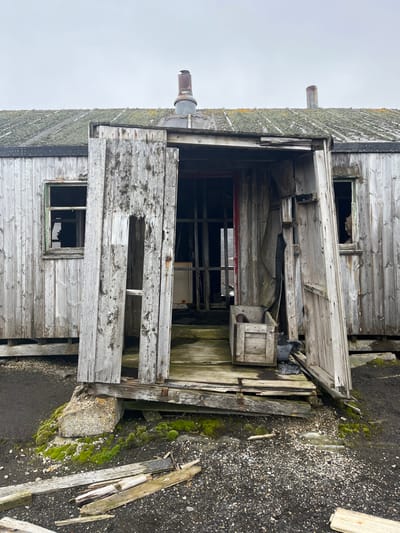 An old, dilapidated wooden building stands against an overcast sky. The weathered structure features broken windows, a partially collapsed doorway, and a chimney pipe extending from the roof. The wood is grayed with age and covered in patches of moss. Debris and loose planks are scattered around the base of the building on an ashphalt surface covered in green moss.
