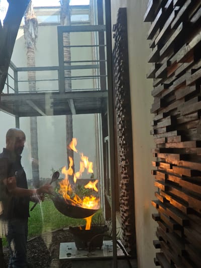 Captured indoors at La Molina, Peru, the scene portrays a man cooking outside, reflected on a glass panel, while the ceiling design above features a series of wooden blocks. Flames are visible, emanating from the cooking vessel he is manipulating.