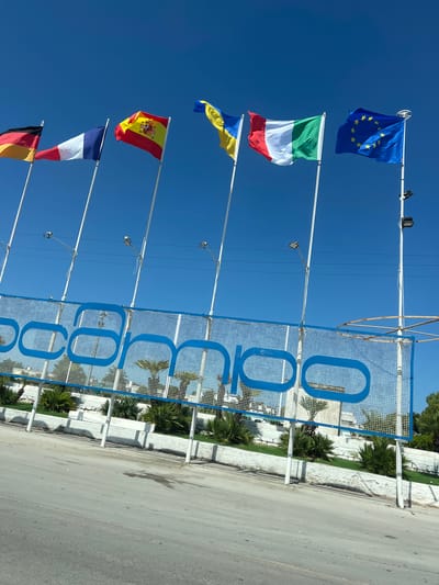 The image depicts an outdoor scene with multiple national flags flying on poles against a clear blue sky. The flags appear to be those of Germany, France, Spain, Ukraine, Italy, and the European Union. Below the flags, there is a large blue sign with white lettering that spells out "CAMPO" across a grid-like fence. The area behind the fence appears to be landscaped with green grass and palm trees. The foreground shows a paved road or walkway. The scene is likely located in Manfredonia, Italy, given the presence of the Italian flag and the context of the sign.