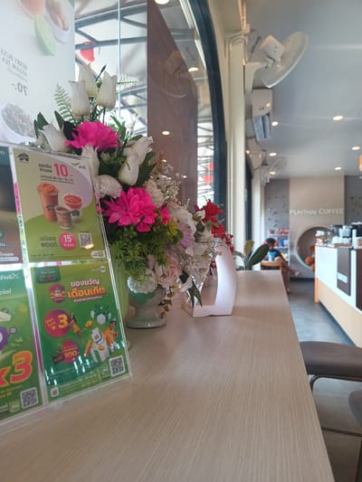 A close-up of a table inside a Punthai Coffee branch in Plai Phraya, Thailand. On the table, there's a flower arrangement in a light green vase, with artificial flowers in shades of white, pink, and red. Next to the flowers are two promotional signs, likely advertising discounts. Behind the flowers is a window, and further into the coffee shop, there are seats, a customer, a counter, and some shelving. There are ceiling fans visible as well. The table is light brown and has a wood grain pattern.