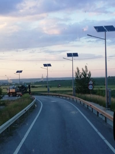 The image depicts a road with a guardrail on the right side, curving to the left. The road surface is asphalt with a white line marking the lane. In the distance, there are fields and some industrial structures. Two solar-powered streetlights are visible, one on the left side of the road and another further down on the right. On the left side of the road, a truck and what appears to be a construction vehicle are parked. The sky is overcast with clouds, suggesting it is either dawn or dusk. There are no people or animals clearly visible in the scene.