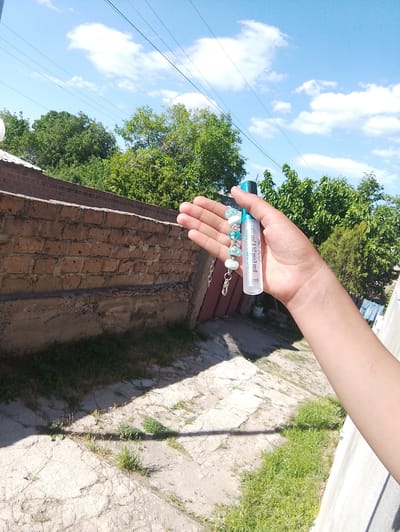 A close-up, waist-up shot shows a person's hand holding a white tube with a blue cap, adorned with a beaded bracelet. The hand and tube are in the foreground, obscuring some of the background. Behind the hand, a brick wall runs horizontally, with patches of grass growing in front of it. Lush green trees fill the mid-ground, with power lines and a bright blue sky with scattered clouds visible in the background. The scene appears to be outdoors, possibly in a residential area, with a clear indication of a sunny day. The image is taken from a slightly upward angle, giving a sense of looking up at the sky and trees. The overall atmosphere is relaxed and casual, suggesting a moment of leisure.