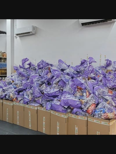 In Bielawska Wieś, Poland, a large pile of gift bags, predominantly purple and clear cellophane, is stacked on top of cardboard boxes. The gift bags are decorated with white snowflake patterns and tied with ribbon. Some bags appear to contain food items, with labels visible on some of the contents. The boxes are sealed with tape that has the "hiper" logo printed repeatedly. In the background, a white wall features two air conditioning units mounted high up. To the left of the gift bags, shelves are visible, stocked with bottles and containers. The scene appears to be indoors, possibly a storage area or a distribution point. There are no people visible in the image.