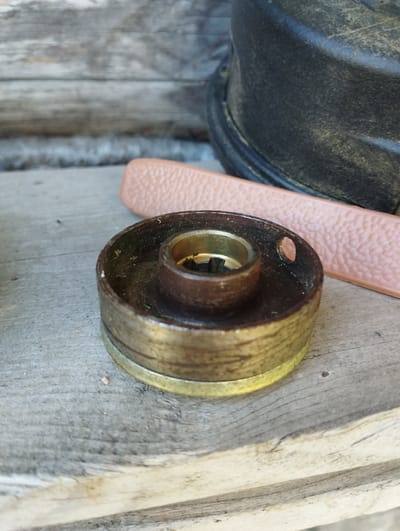 A close-up, high-angle shot captures a worn, brass-colored cylindrical object with a central brass hole on a weathered wooden surface. The object appears to be a pulley or a bearing, with visible layers and a slightly tarnished finish. To the right, a pale pink, textured strap or belt is partially visible, lying on the same wooden surface. In the background, out of focus, is the dark, curved edge of a black, possibly plastic object. The overall scene suggests an outdoor or workshop environment, given the rustic wooden texture and the weathered appearance of the objects. The location is specified as Ширяево (ильинский), Russia, though no specific landmarks or context from the city are discernible within the frame.