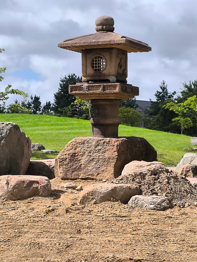 The image shows a Japanese stone lantern placed on a bed of gravel and rocks. The lantern is made of weathered stone and features a pagoda-style roof and a cylindrical body with a square opening containing a grid pattern. It is surrounded by large, rough-hewn rocks. In the background, a lush green hill slopes upwards, dotted with trees and a building with a dark roof. The sky above is partly cloudy. The scene appears to be an outdoor garden or park setting. No people are visible. The location in Mažučiai, Lithuania, is not discernable from the image itself.