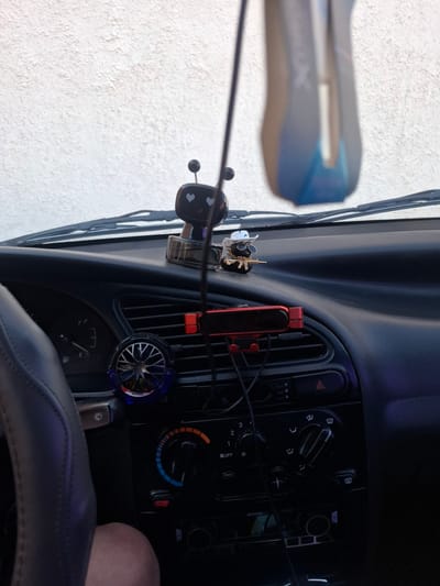 The image depicts the interior of a car's dashboard. In the foreground, a dark-colored steering wheel is visible on the left. The dashboard itself is black plastic, featuring various controls and displays. A fuel gauge is present on the left side. In the center of the dashboard, an air conditioning vent is visible, with a red phone mount attached to it. Below the vent are climate control knobs and buttons, including temperature and fan speed controls. A small, blue fan with a warning label is attached to another vent. On the dashboard above the vents, a small figurine of a character with heart-shaped eyes and an antenna is placed. Further to the right, another figurine of a black character wearing a white cowboy hat and sunglasses, holding a golden gun, is positioned. The car appears to be parked outdoors, with a textured white wall visible through the windshield in the background, illuminated by natural light. The overall scene is that of a car interior with decorative items and operational controls.