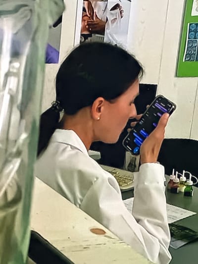 The image shows a woman wearing a white lab coat, with her dark hair pulled back in a ponytail. She is looking at a smartphone held in her right hand. The phone screen displays a graphic with purple and blue bars, resembling a waveform or data visualization. The woman is wearing a small, gold hoop earring in her visible ear. She is seated, and her left hand rests on her lap. In the background, other elements are visible, including a poster or chart with multiple square images on a green background, and what appears to be a laboratory or medical setting with bottles and papers on a table. The scene suggests a professional environment.