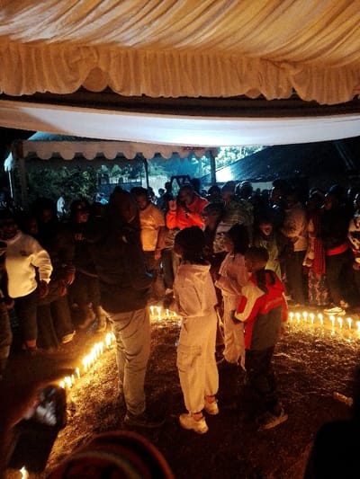 The image depicts a nighttime outdoor gathering under white and cream fabric canopies in Marenyo-Shianda ward, Kenya. A large group of people, including adults and children, are present. The ground, appearing to be dirt or sparse vegetation, is illuminated by numerous small white candles arranged in a curved pattern in the foreground and midground. Overhead, additional ambient lighting illuminates the canopies.
In the center foreground, a tall individual in a dark jacket and light pants stands with their back partially to the camera. To their right, a person in a white hooded jumpsuit and white sneakers stands, facing away. A young child in a red and dark jacket stands next to them, holding onto the person in white. In the midground, a crowd is visible, with a person in a bright orange jacket and dark pants prominent on the right, appearing animated with a raised hand. Other individuals are wearing various casual attire. Many faces are partially obscured by the low light and crowd density.
In the upper midground, a smaller tent structure with scalloped edges is visible, displaying the partial word "AOLANDA." Trees and dark foliage are visible beyond the tent structures, indicating an outdoor, natural environment. In the lower left corner, a hand holds a mobile phone, actively pointing it towards the scene, suggesting photography or videography is occurring.