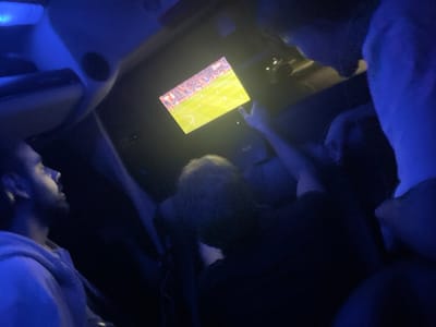 Three people in a dimly lit vehicle watch a soccer match on a screen mounted in the front. The illumination is blue. One person in a light hoodie looks up at the screen. Another person is in the center, pointing at the screen with their right hand. The third person is on the right side of the frame.