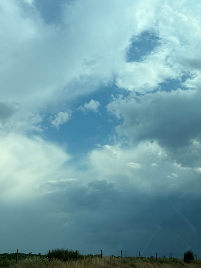 This media file depicts a cloudy sky over a rural landscape. The sky is filled with various types of clouds, ranging from thin, wispy cirrus clouds to larger, more voluminous cumulus clouds. Some areas show patches of bright blue sky peeking through. On the right side of the image, a fence line runs horizontally, bordering a dry, grassy field with some scattered vegetation and a few trees in the distance. The fence appears to be made of wooden posts and wire. The overall impression is of an outdoor scene, likely during the daytime, with overcast or partly cloudy weather. There is no visible text in the image. The scene evokes a sense of natural openness and perhaps a touch of pastoral tranquility. The orientation of the image is such that the sky occupies the majority of the frame, with the ground and fence line forming a lower border on the right.