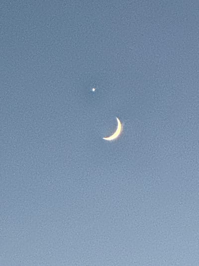 The image displays a clear, pale blue sky with a very faint texture of noise. In the upper half of the frame, there is a small, bright white point of light, possibly a star or planet. To the lower right of this point of light, a thin, crescent moon is visible, illuminated by a warm, yellow light. The moon is curved and positioned diagonally within the frame. The image is a close-up shot of the sky, with no discernible urban or natural landscape elements other than the celestial bodies. The location in Le Brassus, Switzerland, cannot be determined from the visual information provided in the image.