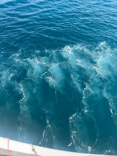 The image shows a view of the sea from the perspective of a boat. The water is a deep blue and appears to be choppy, with white foam and streaks created by the boat's movement. The line where the calm water meets the disturbed water is visible. In the foreground, a portion of the white railing of the boat is visible, with red and yellow ropes attached. The location is the Municipal Unit of Kastro - Kyllini, Greece. The scene depicts a marine environment in motion.