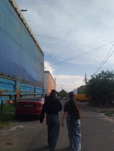 In Nor Geghi, Armenia, two young women walk down an asphalt road, viewed from behind. The woman on the left wears a dark, long-sleeved top and loose-fitting blue jeans. The woman on the right wears a brown short-sleeved top and similar loose-fitting blue jeans. They are walking away from the camera, with their right arms extended.

To their left, a large, tall, blue truck trailer is parked, showing rust on its lower panels. In front of the truck, a red Hyundai Elantra sedan is parked, with its license plate visible: "AM 328 CQ 61".

Further down the road, an orange truck trailer is parked. Beyond that, a yellow van is visible. The background features a tree on the right side of the road and a clear sky with scattered clouds above. A few people are visible near a red bicycle further down the road. The scene suggests a residential or industrial area, possibly on the outskirts of the village.